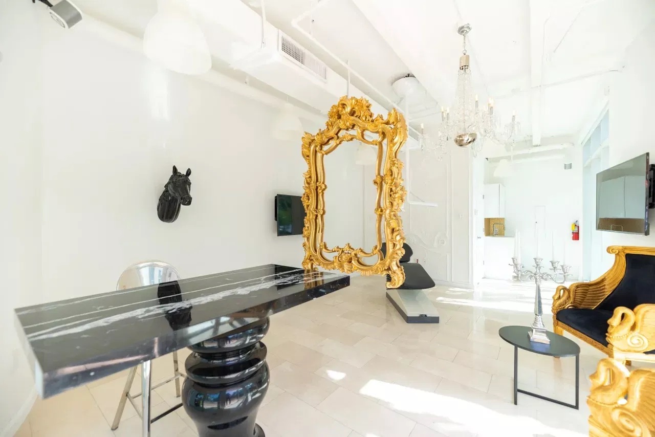 The image shows an interior view of a modern living space with luxurious furnishings, including a large mirror, chandeliers, and decorative items, set against white walls and a black floor.