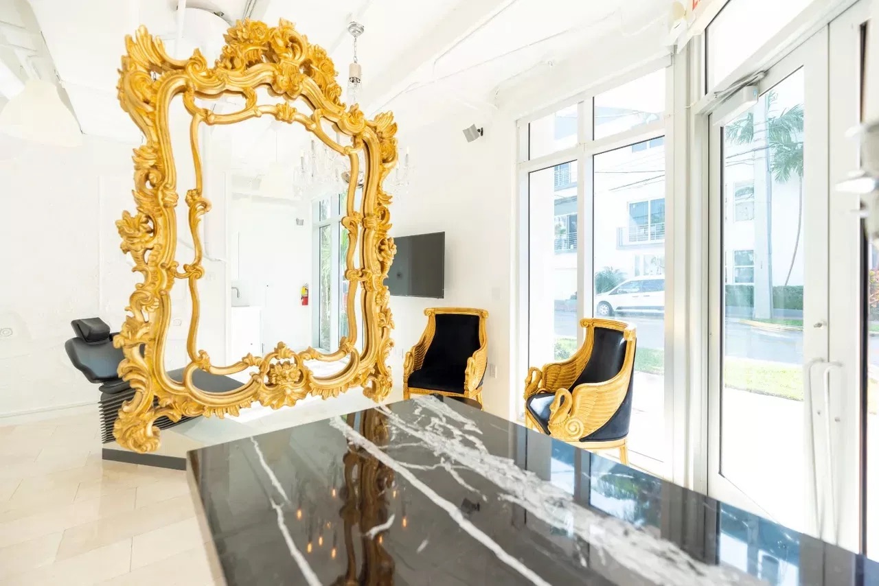 The image depicts a luxurious interior space with an extravagant gold-framed mirror mounted on a wall above a black table, accompanied by two chairs, set against a large window allowing natural light to enter the room.