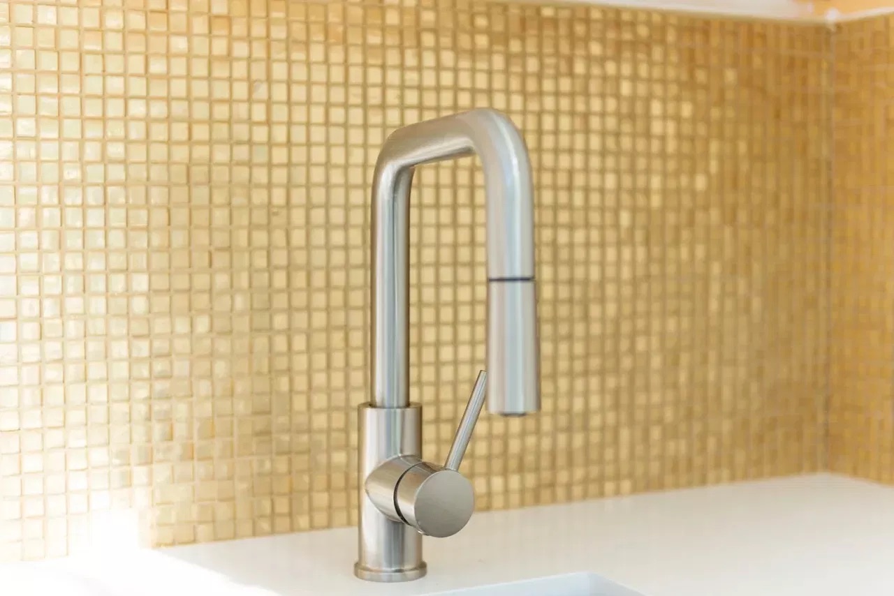 The image shows a modern kitchen faucet with a high-arc design mounted on a backsplash featuring gold-colored tiles, positioned above a white countertop.