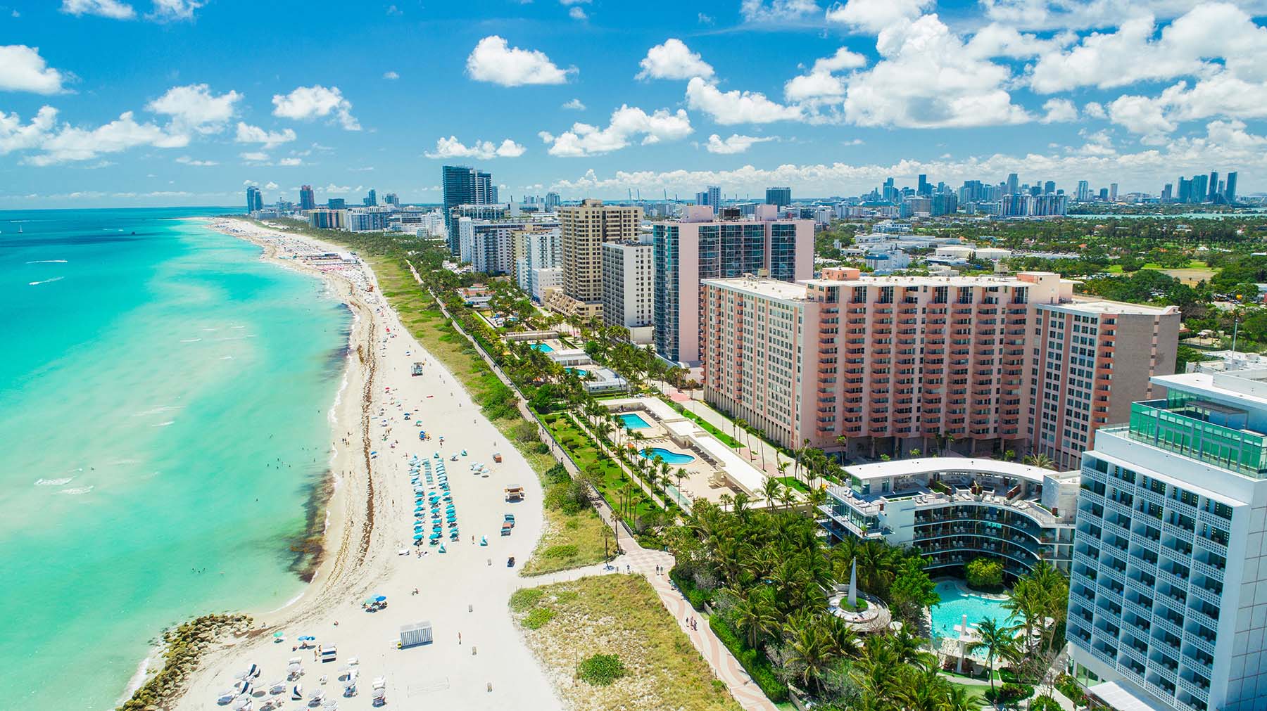 A vibrant beach scene with clear blue waters, white sandy beaches, palm trees, resort buildings, and a city skyline in the background under a bright sunny day.