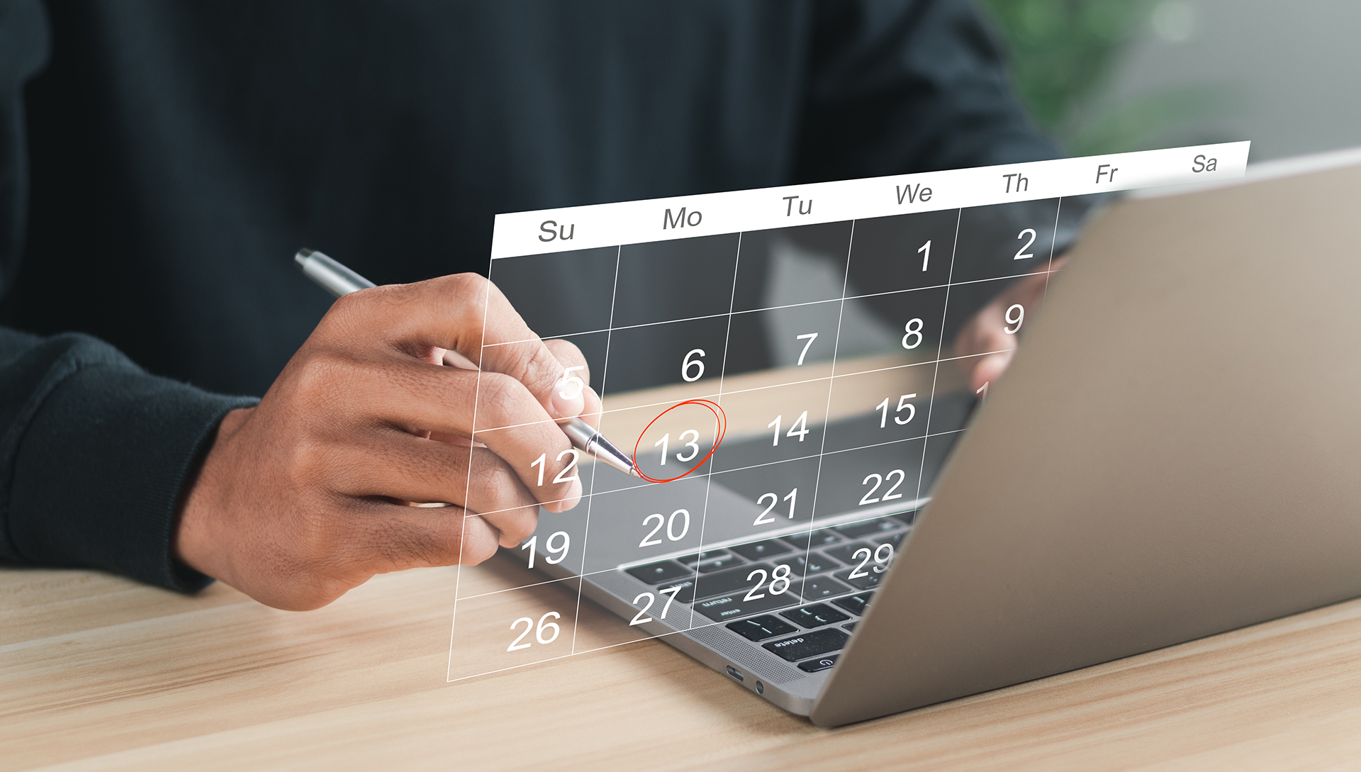 The image shows a person s hands interacting with a digital calendar displayed on a laptop screen, which is placed on a wooden table.