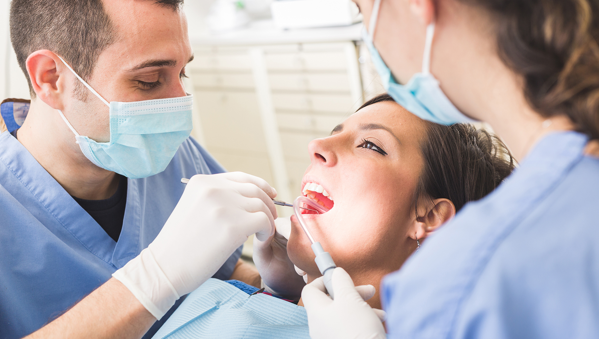 The image shows a dental professional performing a procedure on a patient, with the dentist using a drill and wearing protective gloves and a mask.