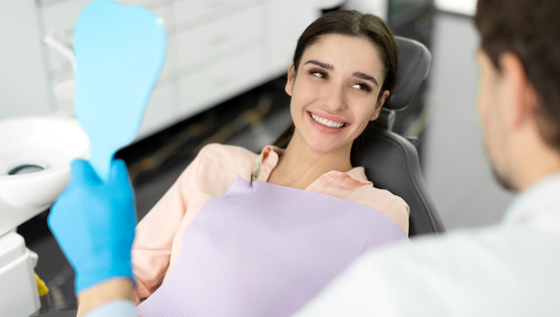 A woman is seated in a dental chair, smiling at the camera, while a dentist works on her teeth.