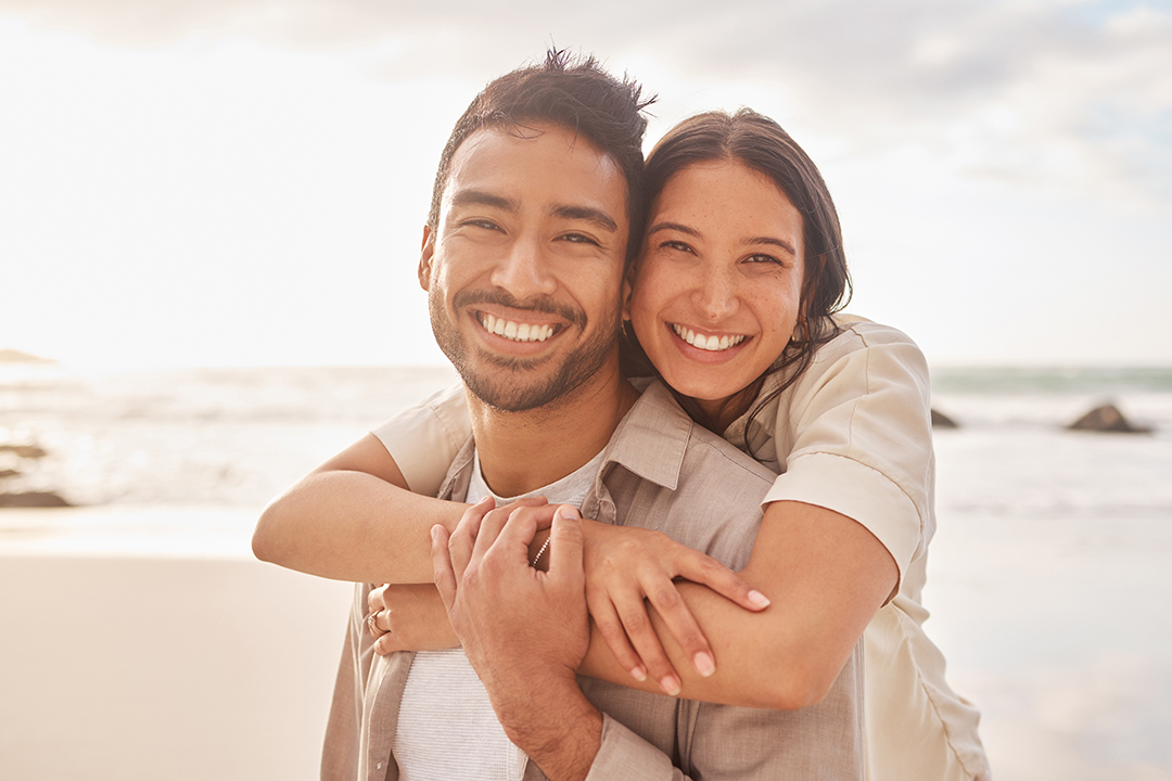 A man and a woman are embracing on a beach, both smiling.