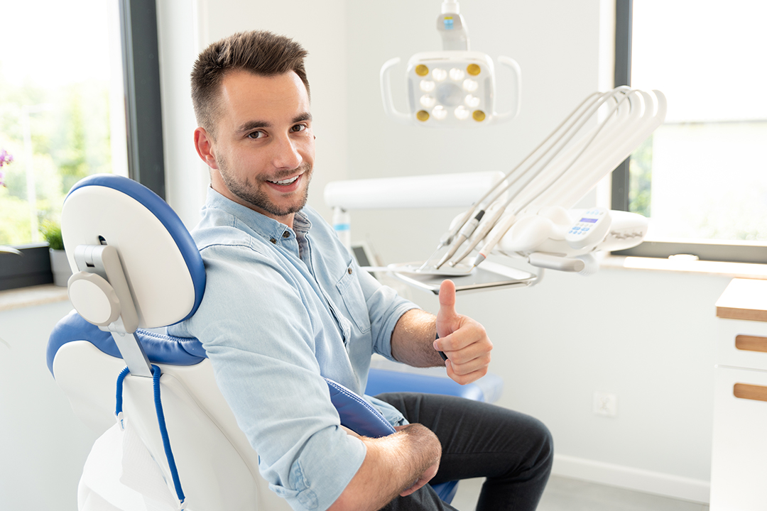 The image shows a man sitting in a dental chair, giving a thumbs-up gesture towards the camera. He is smiling and appears to be in a dental office setting.
