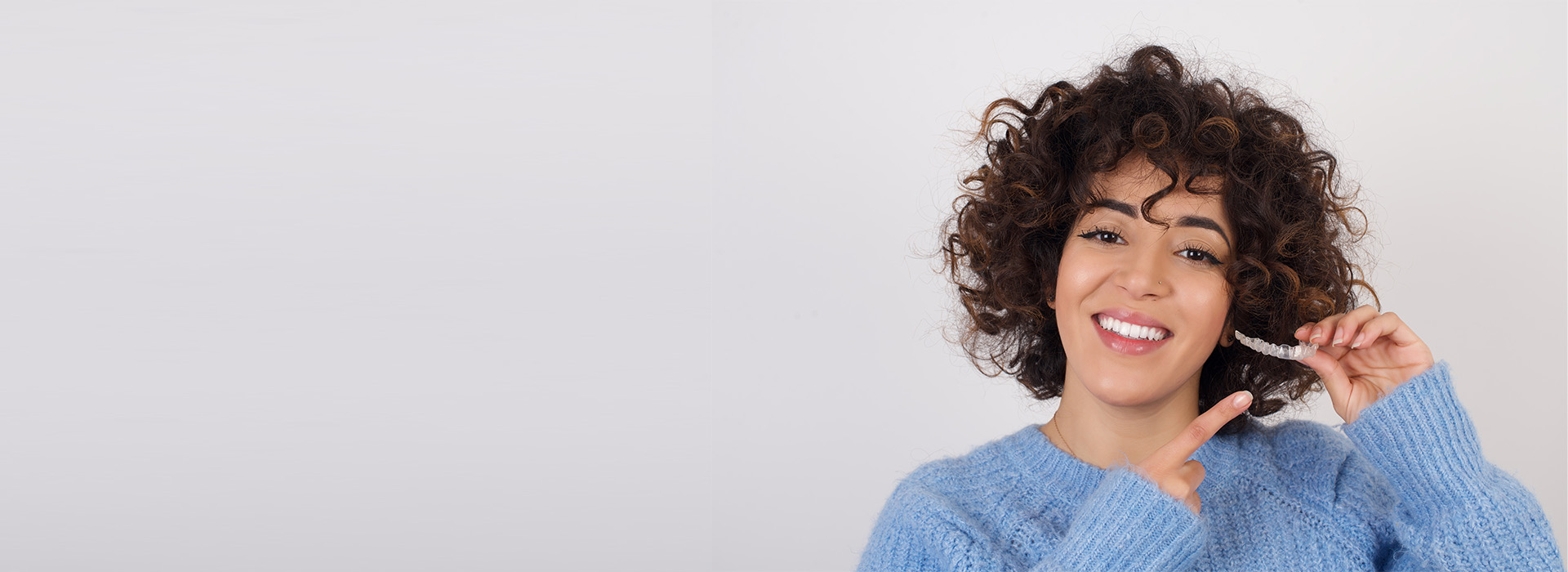 The image shows a person smiling at the camera, with their hair styled in curls, wearing a blue top and holding what appears to be a mobile phone. They are set against a plain background.