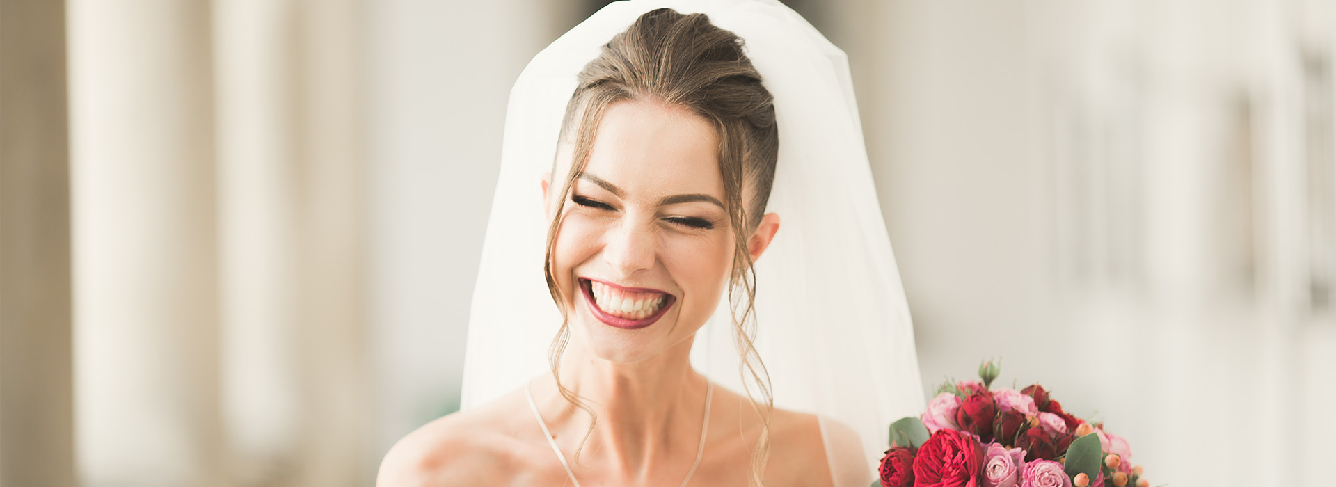 A bride in a white veil and dress, smiling and laughing while holding a bouquet of flowers.