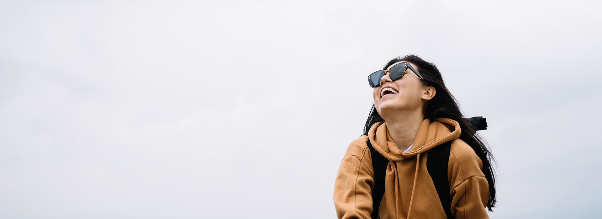 An individual wearing sunglasses and a backpack, smiling at the camera against a cloudy sky background.