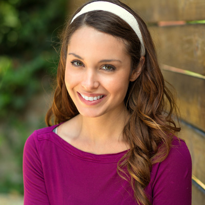 A young woman with long brown hair, wearing a purple top and headband, smiling at the camera.
