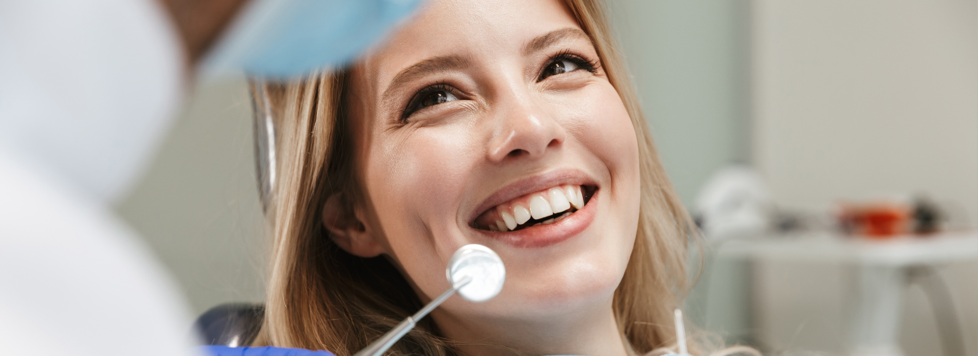 The image shows a woman with a radiant smile, sitting in a dental chair, being attended to by a dental professional who appears to be administering care or treatment.