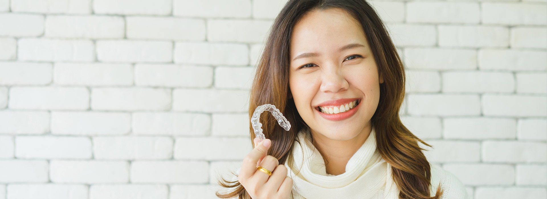 A smiling woman holding a ring in front of her face, set against a white brick wall.