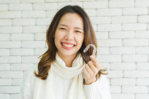 The image shows a woman smiling at the camera while holding an object with the letter S on it, standing against a brick wall background.