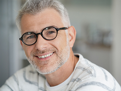 The image shows a man with glasses, smiling at the camera. He has short gray hair and is wearing a white shirt with a patterned collar. The background is blurred but appears to be an indoor setting with natural light.