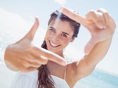 A woman with her hand forming a peace sign, smiling and standing by the ocean under a bright blue sky.